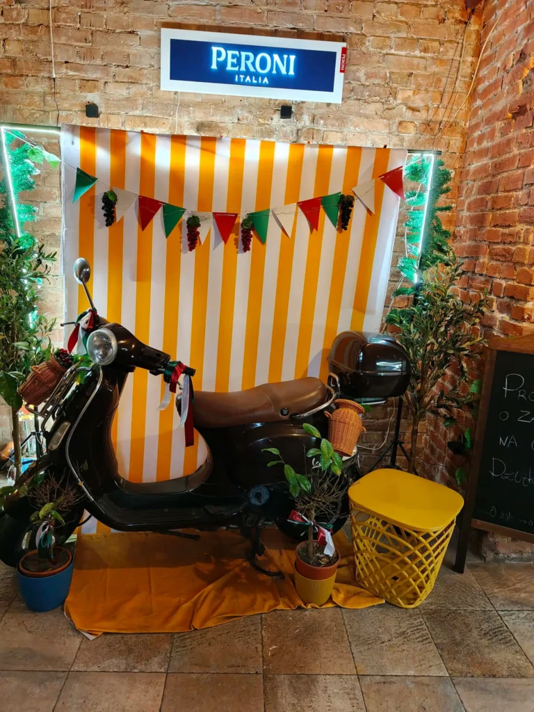 Czarny skuter Vespa na tle pasiastej ścianki pod szyldem Peroni w Restauracja O Sole Mio Italiano