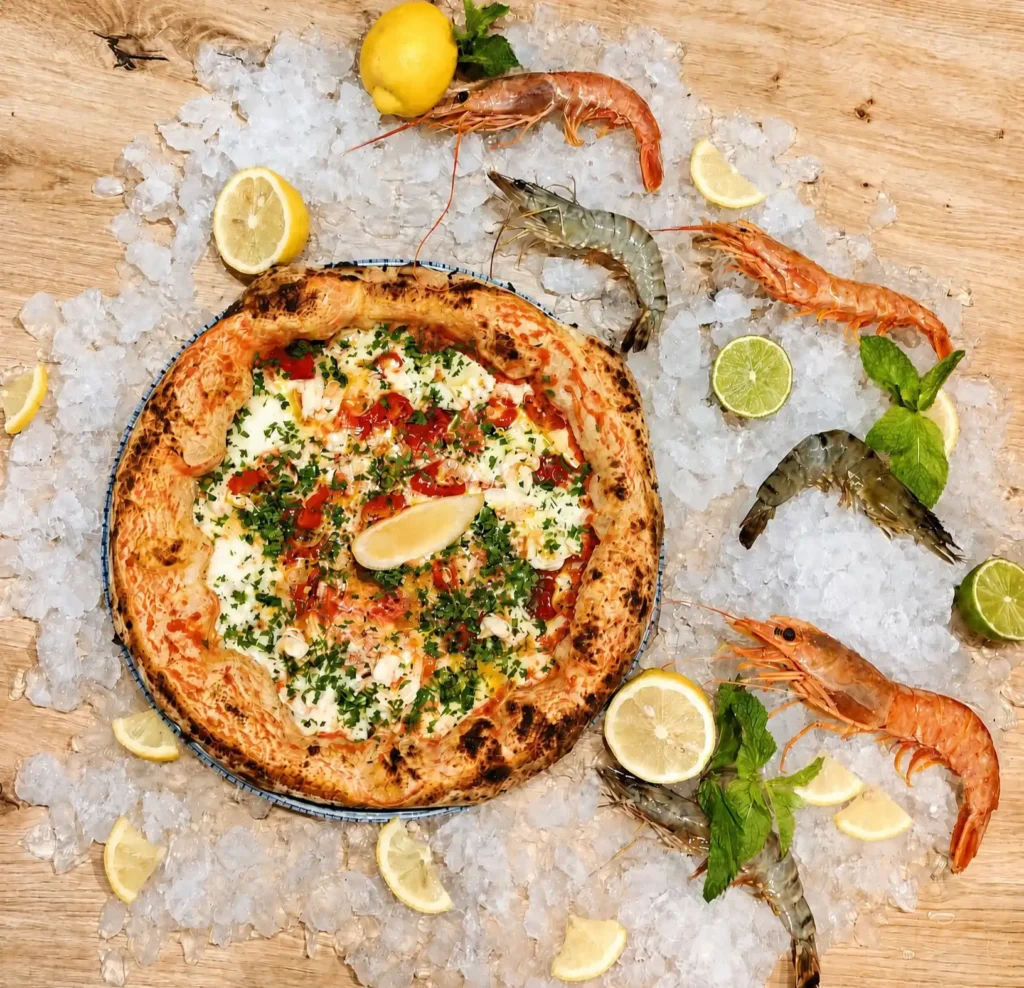 Pizza Diavola di Mare w stylu współczesnej neapolitańskiej — wysoki, wyrośnięty cornicione, krewetki, mozzarella, papryczki chili i świeże zioła, wykończone cytryną. Zdjęcie prezentacyjne na kruszonym lodzie ze świeżymi krewetkami, limonką i miętą.