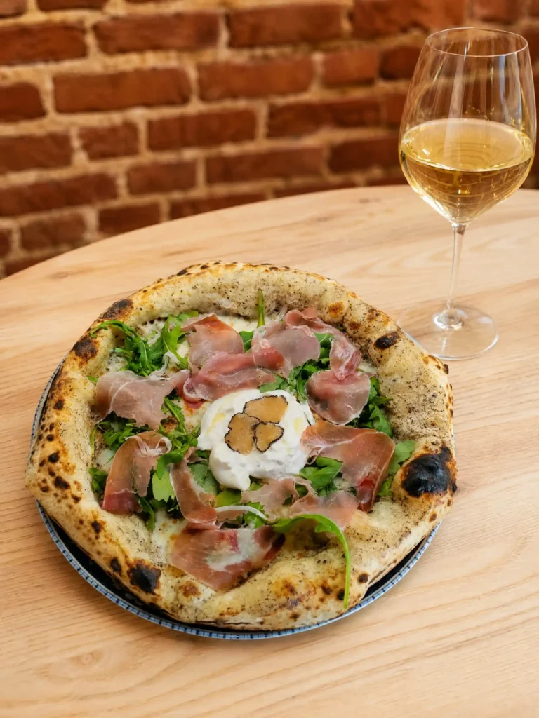 Pizza z prosciutto di Parma, burratą, czarną truflą i rukolą, obok kieliszek białego wina